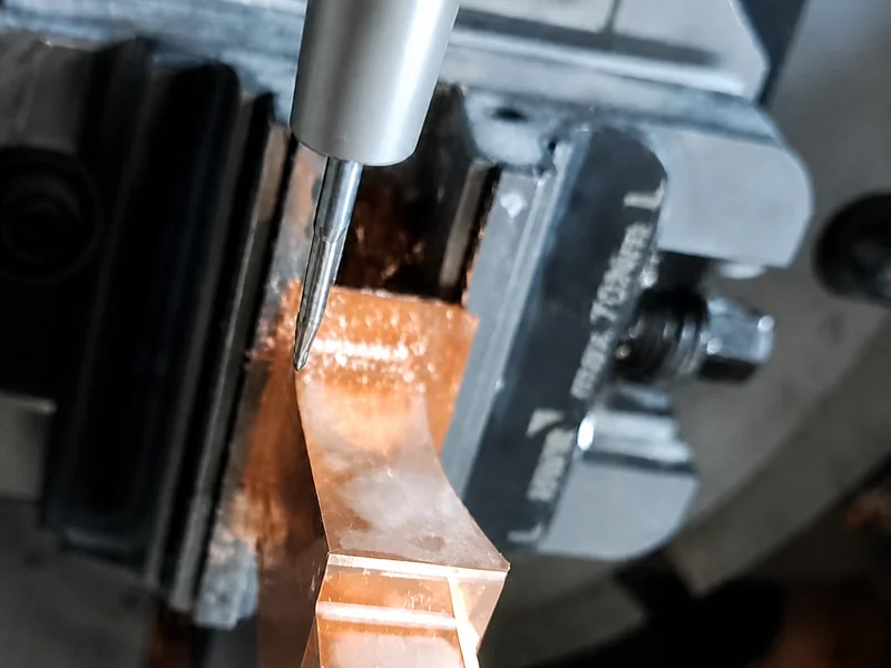 Slender cutting tool above a copper-colored workpiece held in a machine vise.